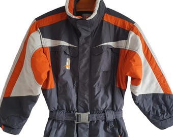 Könnte beinhalten: Grau-oranger Schneeanzug mit weißen Akzenten. Der Schneeanzug hat einen hohen Kragen, einen Gürtel mit silberner Schnalle und Taschen. Die Ärmel und Schultern haben orangefarbene und weiße Einsätze. Geeignet für Winteraktivitäten.