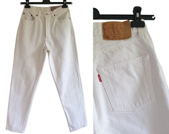 May include: White denim jeans with a classic five-pocket design, displayed on a black hanger. The image shows the front and back, including a small red tag and a brown leather patch.