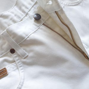 May include: Close-up of white denim jeans with a partially unzipped brass zipper. The jeans feature a button closure, belt loops, and a small brown leather tag with the brand name. The fabric appears to be a sturdy cotton.