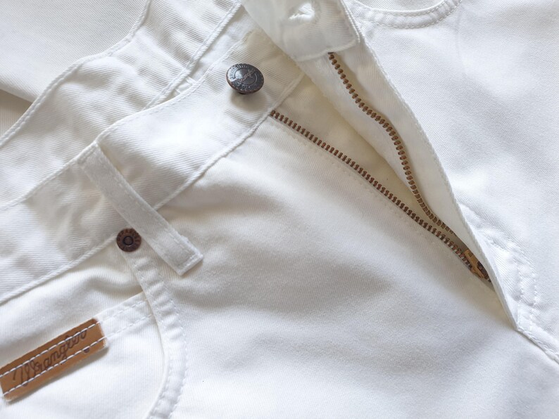 May include: Close-up of white denim jeans with a partially unzipped brass zipper. The jeans feature a button closure, belt loops, and a small brown leather tag with the brand name. The fabric appears to be a sturdy cotton.