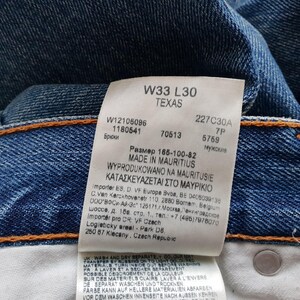 May include: Close-up of blue denim jeans with a white tag. The tag displays the size W33 L30, the word "TEXAS", and other details. The jeans have orange stitching.