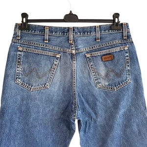 Puede incluir: Vaqueros de mezclilla azul con corte de cintura alta, trabillas para cintur&oacute;n, dos bolsillos traseros con costuras en forma de W y un parche de cuero marr&oacute;n de la marca Wrangler en el bolsillo derecho. La tela tiene un acabado desgastado.