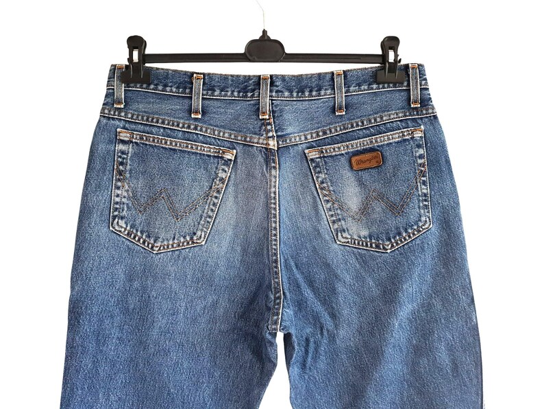 Puede incluir: Vaqueros de mezclilla azul con corte de cintura alta, trabillas para cintur&oacute;n, dos bolsillos traseros con costuras en forma de W y un parche de cuero marr&oacute;n de la marca Wrangler en el bolsillo derecho. La tela tiene un acabado desgastado.