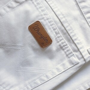 May include: Close-up of white denim jeans with a brown leather Wrangler brand patch on the back pocket. The stitching is visible, and the fabric appears clean and crisp. The jeans are a classic style.