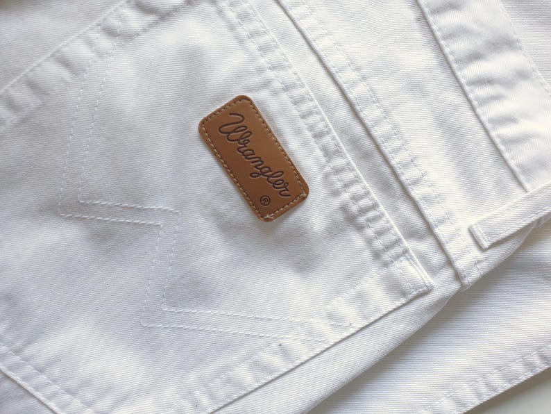 May include: Close-up of white denim jeans with a brown leather Wrangler brand patch on the back pocket. The stitching is visible, and the fabric appears clean and crisp. The jeans are a classic style.
