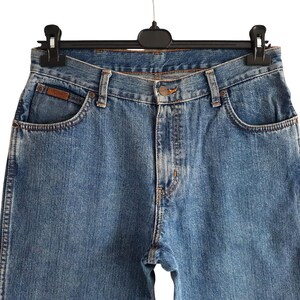 May include: Close-up of blue denim jeans hanging on a black hanger. The jeans have a classic five-pocket design, a button closure, and a brown leather patch on the front pocket. The denim has a faded wash.