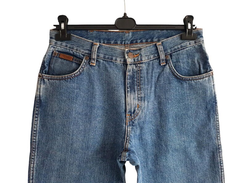 May include: Close-up of blue denim jeans hanging on a black hanger. The jeans have a classic five-pocket design, a button closure, and a brown leather patch on the front pocket. The denim has a faded wash.