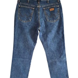 Op de afbeelding: Blauwe denim jeans met het Wrangler-logo op de achterzak, hangend aan een zwarte hanger. De jeans hebben een klassiek ontwerp met vijf zakken en een rechte pijp. De jeans zijn een medium wassing.