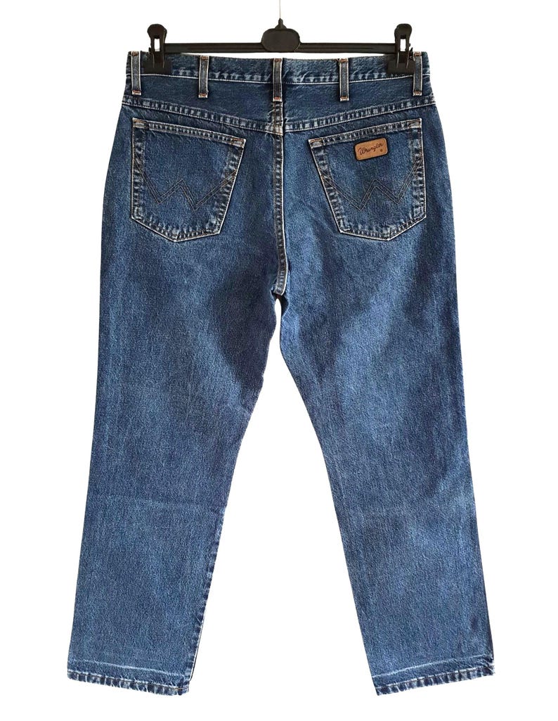 Op de afbeelding: Blauwe denim jeans met het Wrangler-logo op de achterzak, hangend aan een zwarte hanger. De jeans hebben een klassiek ontwerp met vijf zakken en een rechte pijp. De jeans zijn een medium wassing.