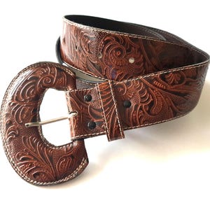 Può includere: Una cintura in pelle marrone con un design floreale ornato e una grande fibbia abbinata. La cintura è cucita con filo bianco.