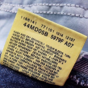 Op de afbeelding: Close-up van een geel kledinglabel met zwarte tekst, bevestigd aan blauwe denimstof. Het label bevat wasvoorschriften en productcodes. Het rechthoekige label heeft afgeronde hoeken.