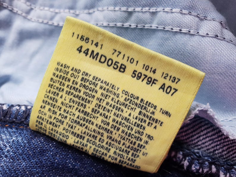 Op de afbeelding: Close-up van een geel kledinglabel met zwarte tekst, bevestigd aan blauwe denimstof. Het label bevat wasvoorschriften en productcodes. Het rechthoekige label heeft afgeronde hoeken.