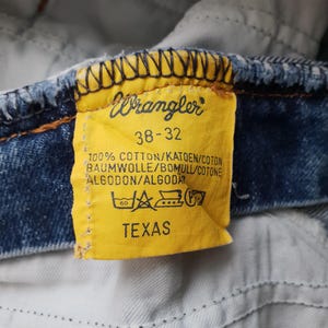 Puede incluir: Una etiqueta de ropa de tela amarilla unida a una tela de mezclilla azul. La etiqueta presenta el logotipo de Wrangler, la talla 38-32, el texto 100% ALGOD&Oacute;N, la palabra TEXAS y varios s&iacute;mbolos de cuidado impresos en tinta negra sobre el fondo.