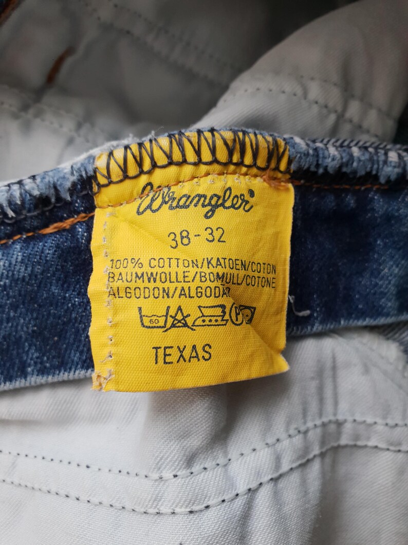 Puede incluir: Una etiqueta de ropa de tela amarilla unida a una tela de mezclilla azul. La etiqueta presenta el logotipo de Wrangler, la talla 38-32, el texto 100% ALGOD&Oacute;N, la palabra TEXAS y varios s&iacute;mbolos de cuidado impresos en tinta negra sobre el fondo.
