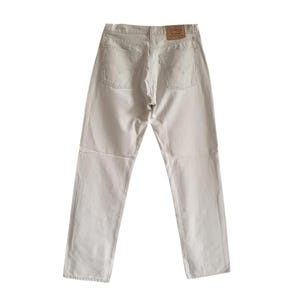 Puede incluir: Vista trasera de unos vaqueros de mezclilla beige claro con un diseño clásico de cinco bolsillos. Los vaqueros tienen un parche de cuero marrón con un logotipo y una pequeña etiqueta roja. Los vaqueros son de color neutro y tienen un corte recto.