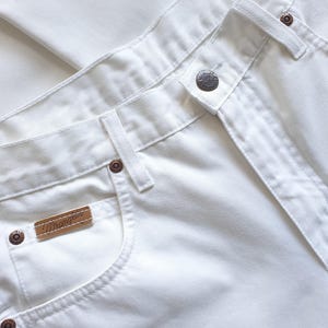 May include: Close-up of white Wrangler jeans. The jeans feature a button closure, belt loops, and a small brown leather tag with the Wrangler logo. The pockets are visible, and the stitching is white. The jeans are made of a sturdy, durable fabric.