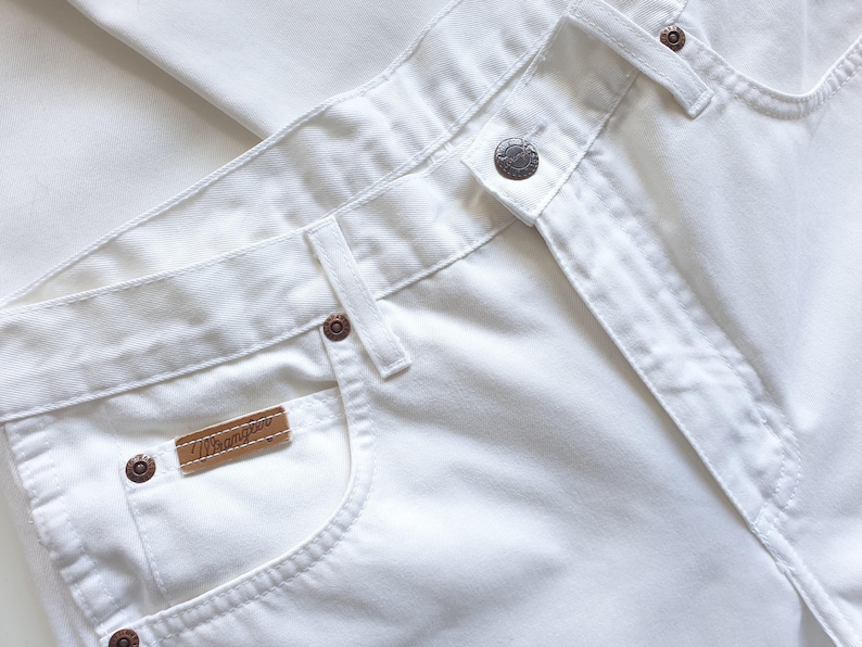 May include: Close-up of white Wrangler jeans. The jeans feature a button closure, belt loops, and a small brown leather tag with the Wrangler logo. The pockets are visible, and the stitching is white. The jeans are made of a sturdy, durable fabric.