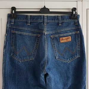 May include: A pair of blue denim jeans with a brown leather Wrangler label on the back pocket.