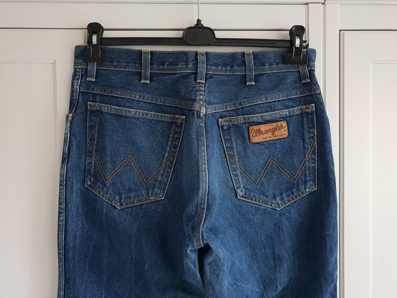 May include: A pair of blue denim jeans with a brown leather Wrangler label on the back pocket.