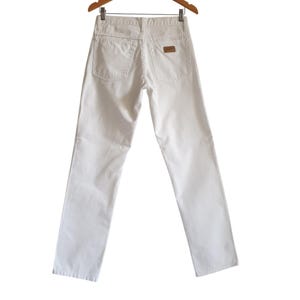 May include: A pair of white denim pants hanging on a wooden hanger. The pants have a classic five-pocket design with a small brown patch on the back pocket. The pants are a solid, bright white color.
