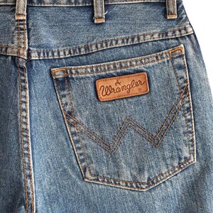 May include: Close-up of blue denim jeans with a brown leather Wrangler patch on the back pocket. The jeans are hanging on a black hanger. The stitching is visible in a contrasting tan color.