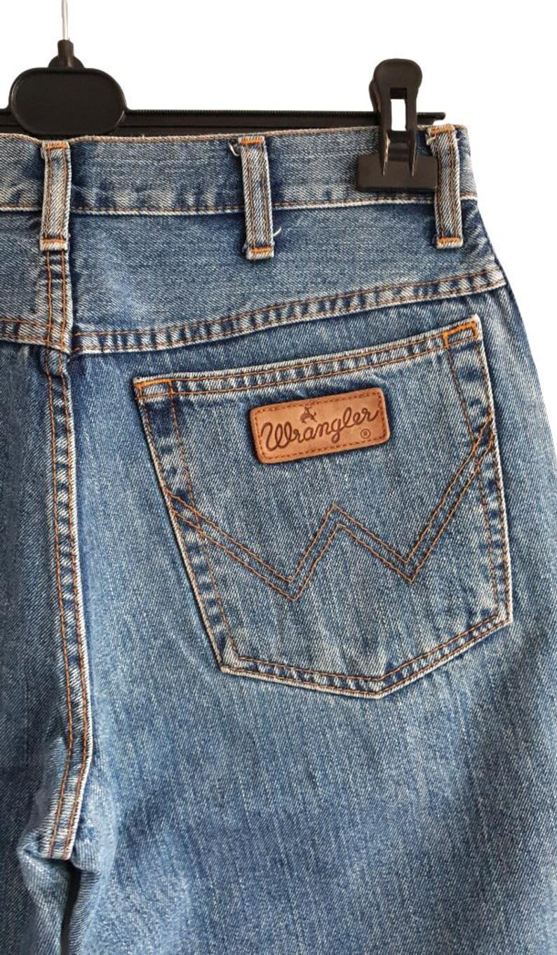 May include: Close-up of blue denim jeans with a brown leather Wrangler patch on the back pocket. The jeans are hanging on a black hanger. The stitching is visible in a contrasting tan color.