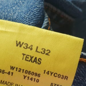 May include: A yellow tag with the text "W34 L32 TEXAS" and additional numbers and letters. The tag is attached to a pair of blue jeans.
