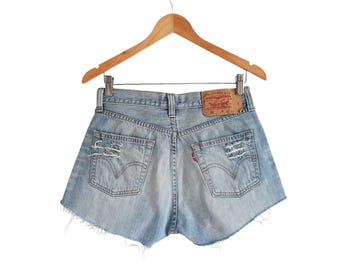 Puede incluir: Pantalones cortos de mezclilla azul claro con bordes deshilachados y un parche marrón de Levi's en el bolsillo trasero. Los pantalones cortos están colgados en una percha de madera.