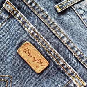 Op de afbeelding: Close-up van blauwe denimstof met oranje stiksels. Een bruin leren label met de merknaam "Wrangler" is op de denim genaaid. De stiksels en het label zijn opvallende kenmerken van de jeans.