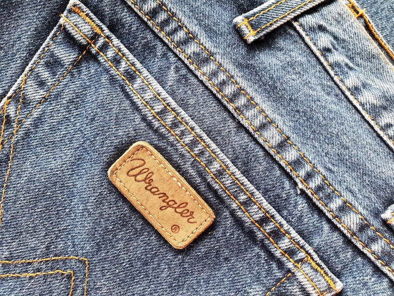 Op de afbeelding: Close-up van blauwe denimstof met oranje stiksels. Een bruin leren label met de merknaam "Wrangler" is op de denim genaaid. De stiksels en het label zijn opvallende kenmerken van de jeans.