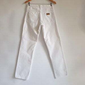May include: White denim jeans hanging on a wooden hanger. The jeans have two back pockets and a small brown leather patch. The pants are straight-legged and appear to be a vintage style.