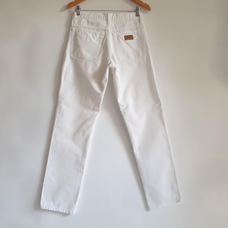 May include: White denim jeans hanging on a wooden hanger. The jeans have two back pockets and a small brown leather patch. The pants are straight-legged and appear to be a vintage style.