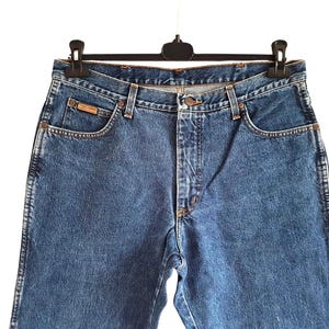 Op de afbeelding: Blauwe spijkerbroek hangend aan een zwarte hanger. De jeans hebben een klassiek ontwerp met vijf zakken, een knoop- en ritssluiting en een klein bruin label op de voorzak. De jeans zijn gemaakt van een medium-wash denim.