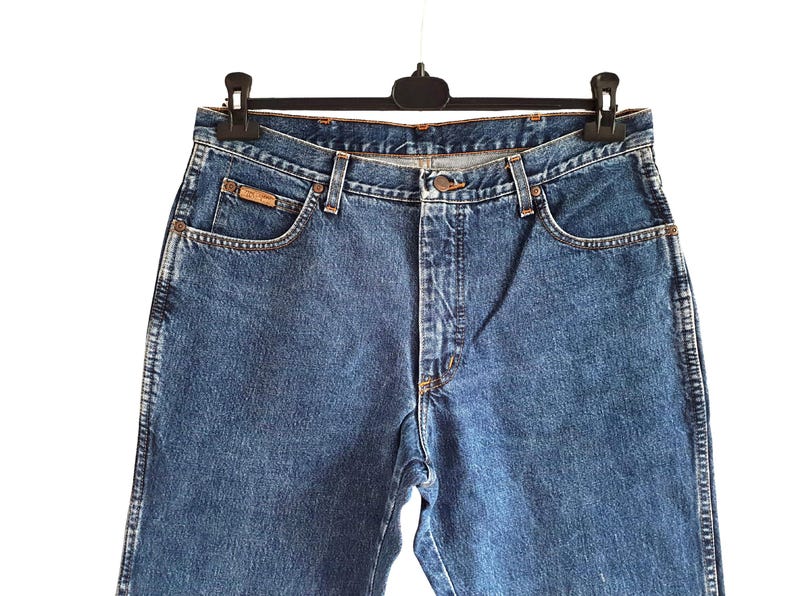 Op de afbeelding: Blauwe spijkerbroek hangend aan een zwarte hanger. De jeans hebben een klassiek ontwerp met vijf zakken, een knoop- en ritssluiting en een klein bruin label op de voorzak. De jeans zijn gemaakt van een medium-wash denim.