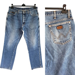 Puede incluir: Vaqueros de mezclilla en azul lavado medio con dise&ntilde;o cl&aacute;sico de cinco bolsillos, cierre de botones y trabillas para cintur&oacute;n. El bolsillo trasero muestra un parche de cuero marr&oacute;n con el logo Wrangler y una costura decorativa en forma de W.