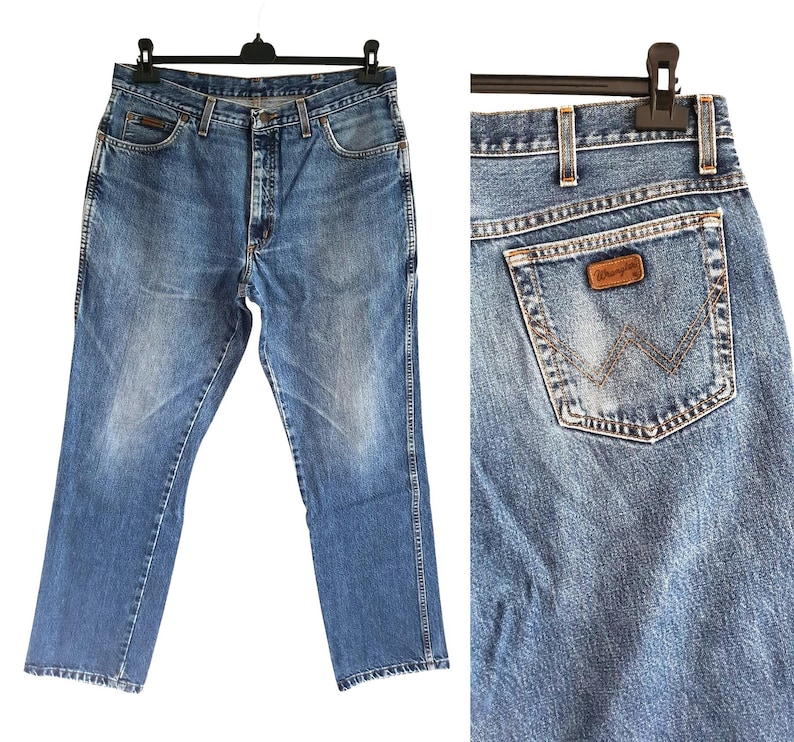 Puede incluir: Vaqueros de mezclilla en azul lavado medio con dise&ntilde;o cl&aacute;sico de cinco bolsillos, cierre de botones y trabillas para cintur&oacute;n. El bolsillo trasero muestra un parche de cuero marr&oacute;n con el logo Wrangler y una costura decorativa en forma de W.