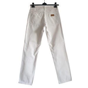 May include: Off-white denim jeans hanging on a black hanger. The jeans have two back pockets and a small brown patch with text on the back. The jeans are straight-legged.