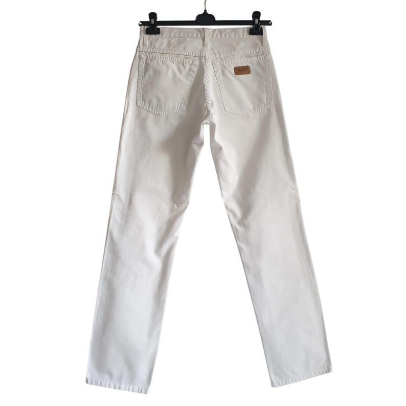 May include: Off-white denim jeans hanging on a black hanger. The jeans have two back pockets and a small brown patch with text on the back. The jeans are straight-legged.
