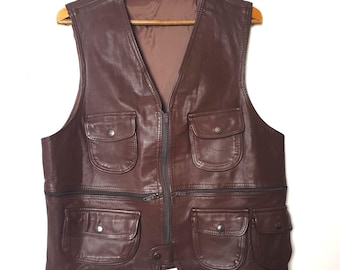 May include: A brown leather vest with a zip-up front and multiple pockets. The vest is hanging on a wooden hanger. The vest has a V-neck and a brown interior lining. The pockets have button closures.