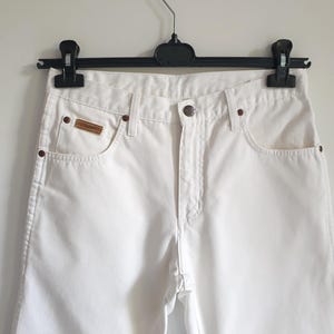 May include: White denim jeans with a classic five-pocket design. The jeans feature a button closure, a small brown leather patch on the front pocket, and copper-colored rivets. The jeans are hanging on a black hanger.