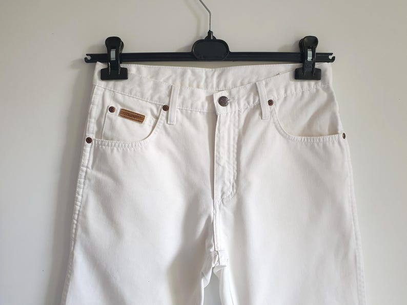 May include: White denim jeans with a classic five-pocket design. The jeans feature a button closure, a small brown leather patch on the front pocket, and copper-colored rivets. The jeans are hanging on a black hanger.