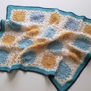 Può includere: Una coperta all'uncinetto con sfondo bianco e quadrati blu e gialli. La coperta ha un bordo turchese.