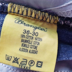 Op de afbeelding: Close-up van een geel Wrangler jeanslabel. Het label toont maat 36-30, stofdetails en wasvoorschriften. Het woord "TEXAS" staat onderaan het label.