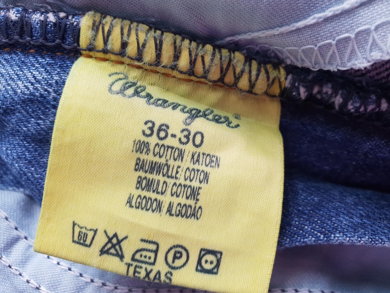 Op de afbeelding: Close-up van een geel Wrangler jeanslabel. Het label toont maat 36-30, stofdetails en wasvoorschriften. Het woord "TEXAS" staat onderaan het label.