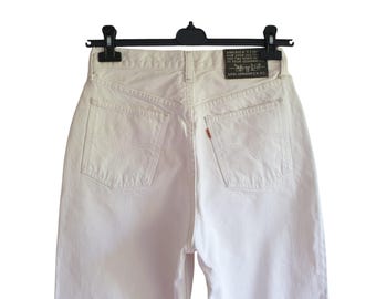 May include: Rear view of white denim jeans suspended from a black hanger. The jeans feature two back pockets with the Levi's logo and a small orange tag. A black leather patch with text is visible.