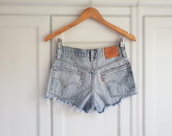 Puede incluir: Pantalones cortos de mezclilla azul claro con bordes deshilachados y un parche de cuero marrón en la parte posterior. Los pantalones cortos están colgados en una percha de madera.