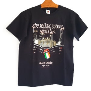Può includere: T-shirt nera con grafica del tour "A Bigger Bang" dei Rolling Stones al Castello di Slane. Il design include il logo della band, un'illustrazione del castello e la data 18 agosto 2007. La maglietta è appesa a una gruccia di legno.