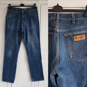 May include: A pair of blue denim jeans with a brown leather Wrangler label on the back pocket. The jeans have a classic straight leg style.