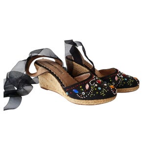 May include: Black wedge espadrille sandals with colorful jewel embellishments. The sandals have a woven jute sole and black ribbon ankle ties. The open-toe shoes feature a scalloped edge and are suitable for casual wear.