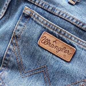 May include: Close-up of blue denim jeans with a brown leather Wrangler patch on the back pocket. The patch features the Wrangler logo in a cursive font. The jeans have orange stitching.